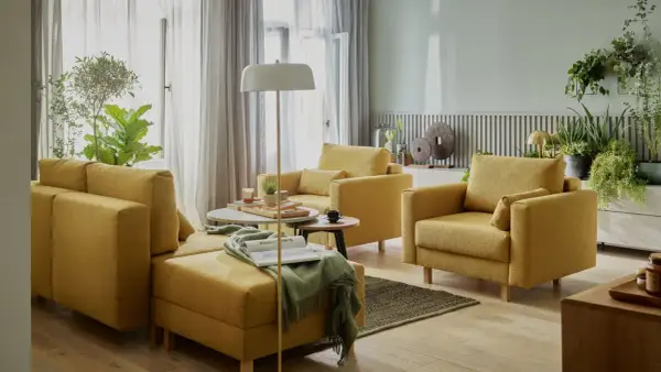 Przytulny salon bez telewizora. Żółte meble wypoczynkowe, stoliki kawowe, lampa podłogowa i mnóstwo zielonych roślin tworzą relaksującą atmosferę.