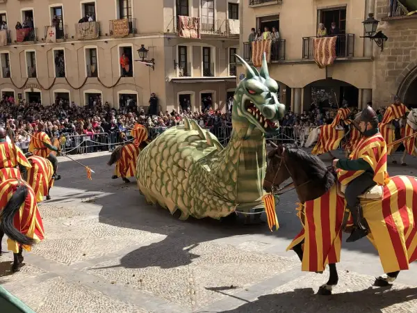 Un dragón gigante desfila en las fiestas de Alcañiz, acompañado por jinetes con trajes de rayas rojas y amarillas.
