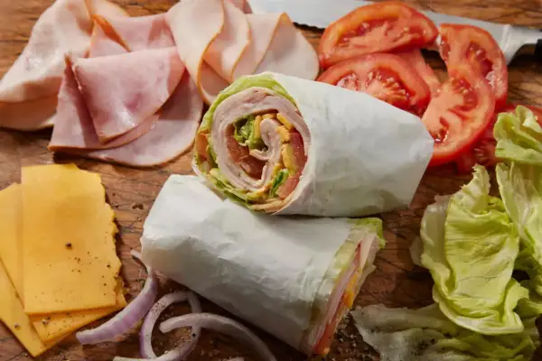 Dwa zawinięte w papier wrapy z szynką, serem, pomidorami i sałatą. Obok plastry szynki, sera, pomidorów i cebuli. Idealne na szybki posiłek, choć warto sprawdzić wrap mcdonald kcal.