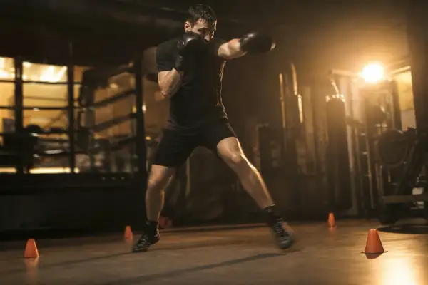 A determined pivot fighter in black boxing gloves and shorts practices in a dimly lit gym, his body a blur of motion.