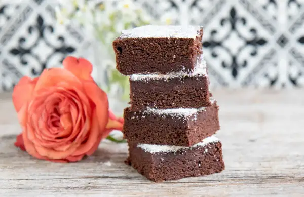 Schokoladige Brownies mit Datteln, bestäubt mit Puderzucker, neben einer zarten orangefarbenen Rose auf Holztisch.