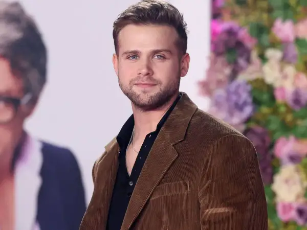 Leo Woodall, con su chaqueta de pana marrón y camisa negra, sonríe ante la cámara.