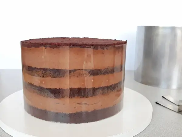 Czekoladowa torta z warstwami ciasta i kremu, przygotowywana w transparentnej formie. Obok stoi metalowy pierścień.