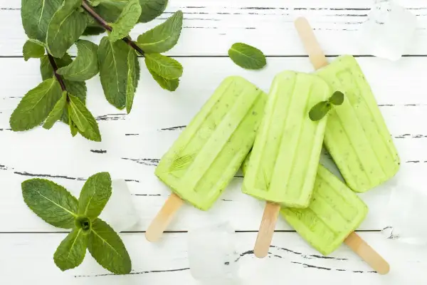 Erfrischendes Minzeis selber machen: Grüne Eis am Stiel mit frischer Minze und Eiswürfeln auf weißem Holzuntergrund.