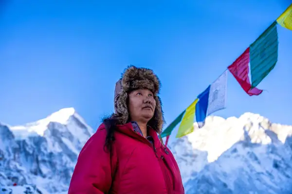 Lhakpa Sherpa, eine Frau mit einem Pelzmützen, blickt in den Himmel, während Gebetsfahnen über schneebedeckten Bergen wehen.