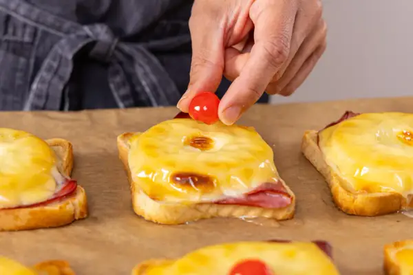 Eine Hand platziert eine Kirsche auf einem überbackenen Toast Hawaii. Der Toast im Backofen ist mit Schinken, Ananas und geschmolzenem Käse belegt.