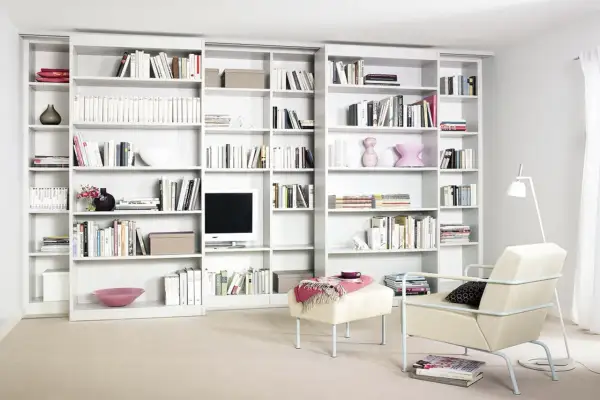 Eine moderne, weiße **eigene Bibliothek** mit vielen Büchern, Dekorationen und einem Fernseher. Ein Sessel und ein Hocker laden zum Verweilen ein.