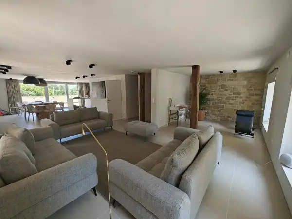 Salon moderne avec canapés gris, un pouf, un tapis, et un mur en pierre apparente rustique. Une plante verte et un fauteuil noir complètent l'espace.