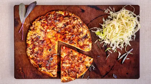 En hel och en skivad pizza på en träskärbräda, med en hög strimlad kål. En klassisk kombination som ger en hint om pizzasallad ursprung.