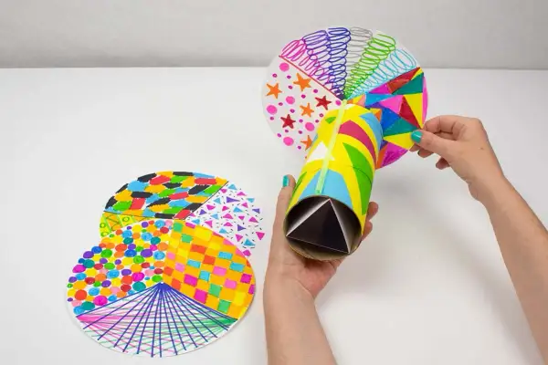 Manos creando un caleidoscopio con manualidades con material reciclado. Tubo de cartón decorado y discos circulares con patrones coloridos.