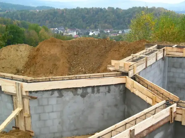 Fundamenty domu w budowie, zbrojenie i szalunki. Widać kopce ziemi i zabudowania w tle. Ważne jest pozwolenie na budowę, by wszystko szło zgodnie z planem.