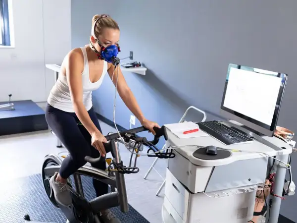 Femme en masque respiratoire effectuant un test d'effort duree sur un vélo d'appartement, connectée à un équipement médical.