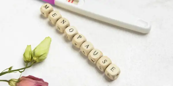 Koraliki z literami układają się w słowo "MENOPAUZA". Obok leży test ciążowy i uschnięte kwiaty, symbolizujące zmiany.