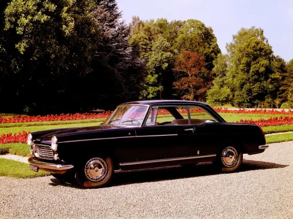 Elegancki, czarny Peugeot 404 coupe z lat 60. stoi na żwirowej alejce, otoczony zielenią i czerwonymi kwiatami.
