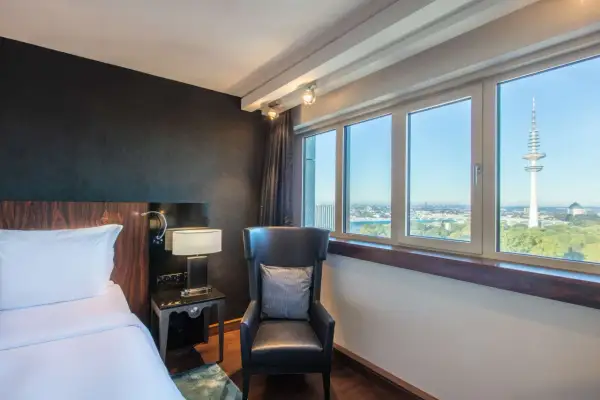 Pokój w Blu Radisson Hamburg z widokiem na wieżę telewizyjną. Wygodne łóżko, fotel i stolik nocny.