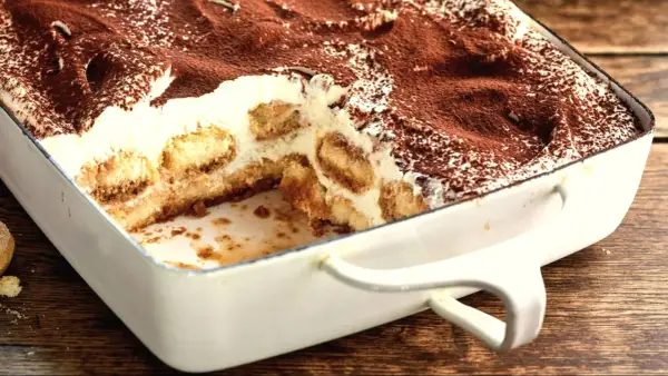 Tiramisu w białej formie, posypane kakao, z widocznymi warstwami biszkoptów i kremu.