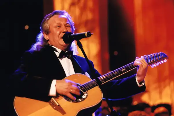 Artysta w czarnym smokingu gra na gitarze, śpiewając do mikrofonu. W tle pomarańczowe światła, jakby na scenie, gdzie kiedyś występowały siedem dziewcząt z Albatrosa.