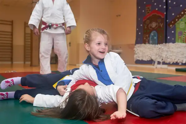 Niñas sonriendo mientras practican judo para niños. Un instructor de judo con cinturón rojo observa.