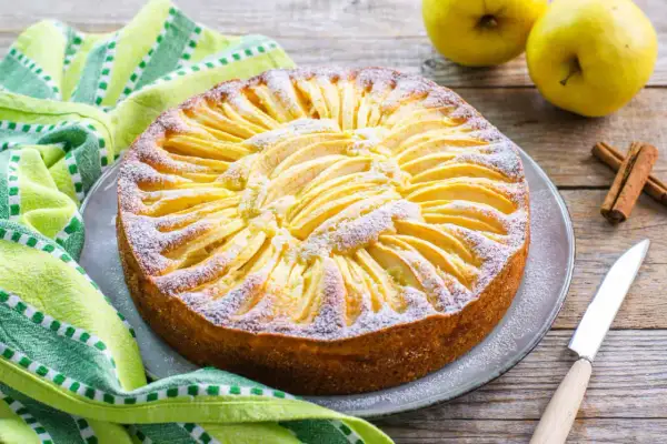 Torta di mele con yogurt, decorata con fettine di mela e zucchero a velo, su un piatto grigio, accanto a un tovagliolo verde e bastoncini di cannella.