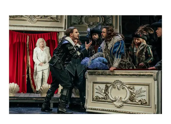 Aktorzy w strojach z epoki na scenie, w tym mężczyzna w czarnym stroju, przypominający Cyrano de Bergerac, gestykuluje do towarzyszy.