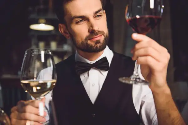 Elegancki mężczyzna w muszce ocenia dwa kieliszki wina. Pokazuje, jak trzymać kieliszek wina, by docenić jego barwę i aromat.