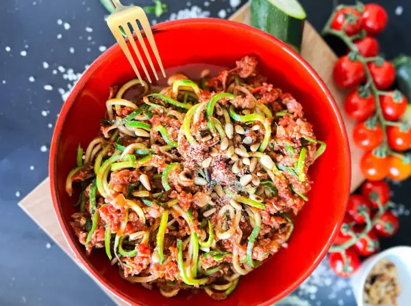 Ein leckeres Zoodles Rezept mit Hackfleischsauce, Pinienkernen und Parmesan in einer roten Schüssel. Daneben Kirschtomaten und Zucchini.