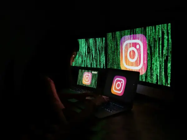 Osoba pracuje przy komputerze, na ekranach widnieje logo Instagrama i zielony kod. Szuka informacji, jak odzyskać zhakowane konto na Instagramie.