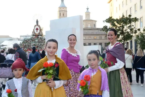 Niñas con trajes regionales y flores rojas, listas para las fiestas Zaragoza. Detrás, un gran tapiz floral y edificios históricos.