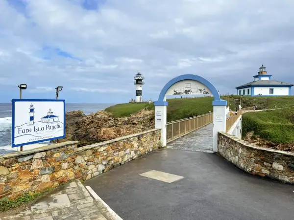 El faro de Ribadeo, con su icónico diseño blanco y negro, se alza sobre un acantilado verde. Un puente arqueado conduce a la isla.