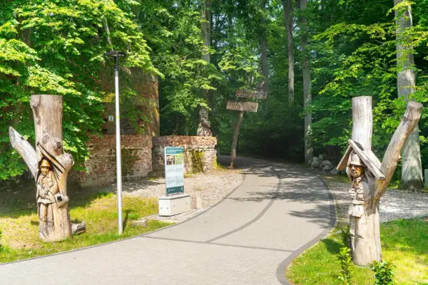 Drewniane rzeźby żołnierzy witają przy ścieżce w lesie. Tablice informacyjne i ruiny ceglane zdradzają, że to Wilczy Szaniec. Ciekawostki czekają!