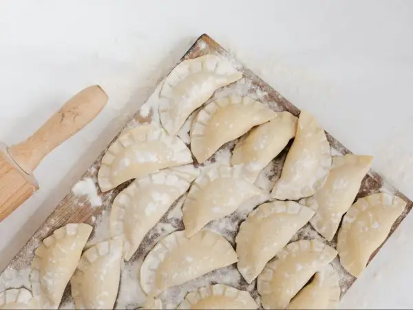 Surowe pierogi ruskie ułożone na desce, obok wałek do ciasta.