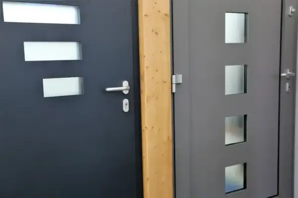 Dwie nowoczesne drzwi, jedna ciemnoszara z trzema prostokątnymi przeszkleniami, druga szara z trzema kwadratowymi, obok drewnianego słupka. Dowiedz się, jak wygłuszyć drzwi.
