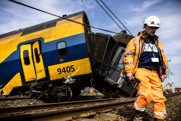 Een ontspoorde trein, nummer 9405, ligt scheef op het spoor. Een man in oranje werkkleding inspecteert de schade, de wagons lijken per ongeluk aan elkaar of los te zijn geraakt.