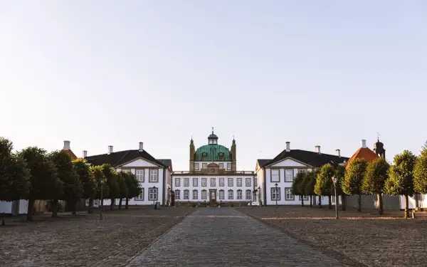 Frederiksborgs slott med sin gröna kupol reser sig majestätiskt mot en klar himmel. En stenlagd väg leder fram till den imponerande byggnaden, flankerad av träd.