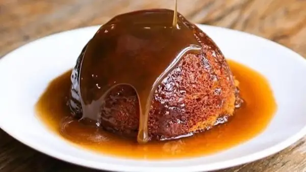 Un delizioso sticky toffee pudding, ricoperto da una cascata di salsa al caramello, servito su un piatto bianco.