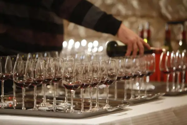 Rzędy kieliszków z czerwonym winem, osoba nalewająca wino z butelki.