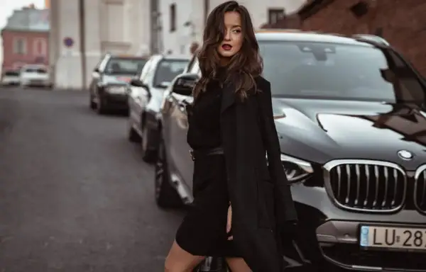 Elegancka kobieta w czarnej sukience swetrowej i płaszczu stoi obok luksusowego BMW na ulicy.