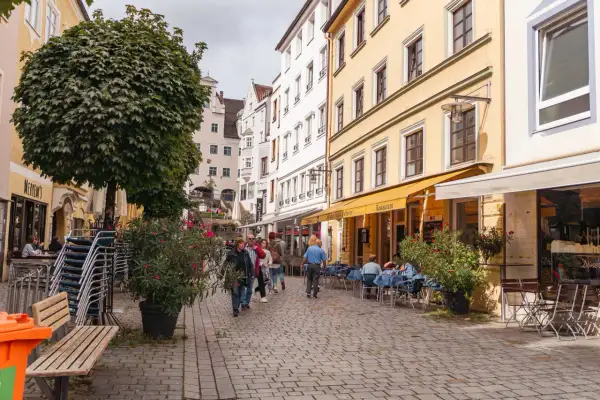 Kempten Geheimtipps: Menschen schlendern durch eine charmante Gasse mit Restaurants und Cafés, umgeben von historischen Gebäuden und üppigem Grün.
