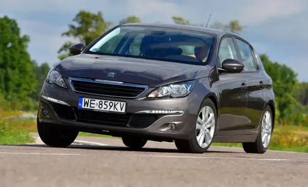 Ciemnoszary Peugeot 308 2015 z polskimi tablicami WE 859KV jedzie po drodze wśród zieleni.