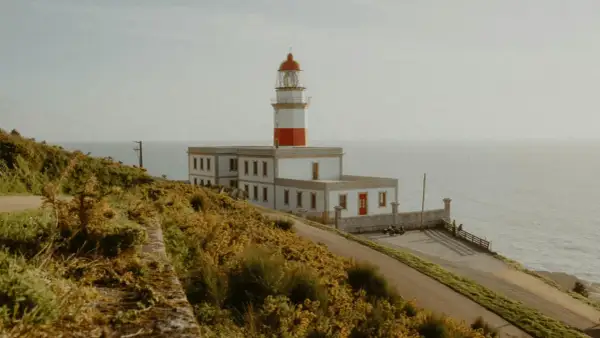El faro de Baiona se alza majestuoso sobre un acantilado cubierto de vegetación dorada, vigilando el mar.
