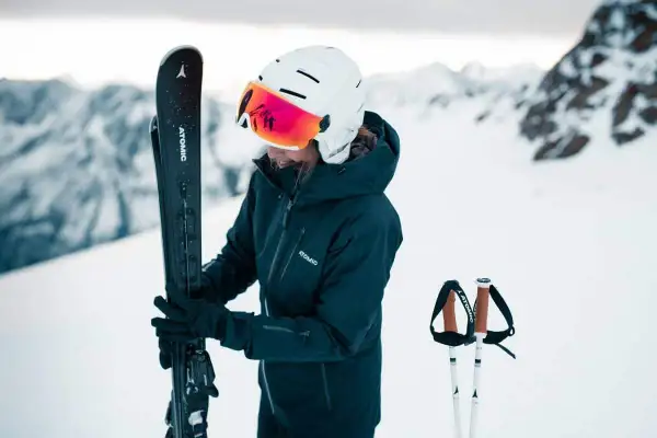 Femme en tenue de ski, tenant ses skis, portant un casque de ski avec visière intégrée.