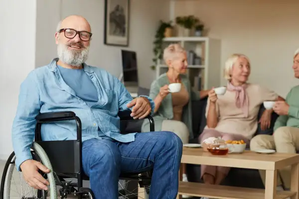 Uomo sorridente in carrozzina, con amici in sottofondo. La sua storia di invalidità civile patologie riconosciute è un esempio di resilienza.