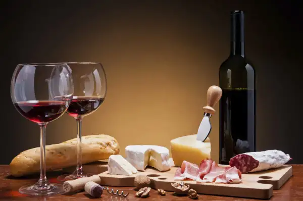 Deux verres de vin rouge, une bouteille, du pain, des fromages, de la charcuterie et des noix. Un accord parfait pour un vin pour fromage et charcuterie.