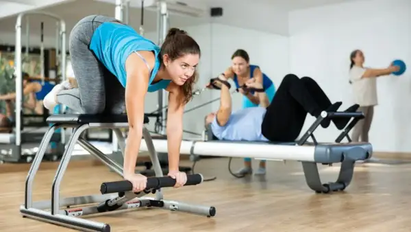 Frauen trainieren im Studio. Eine macht eine Übung auf einem Gerät, während andere im Hintergrund an Pilates-Geräten arbeiten. Das ist Zirkeltraining.