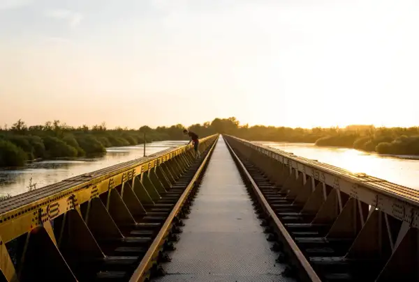 Een man leunt over een spoorbrug bij zonsondergang. De gouden gloed verlicht de rivier en de weelderige vegetatie, wat zorgt voor mooie foto locaties.