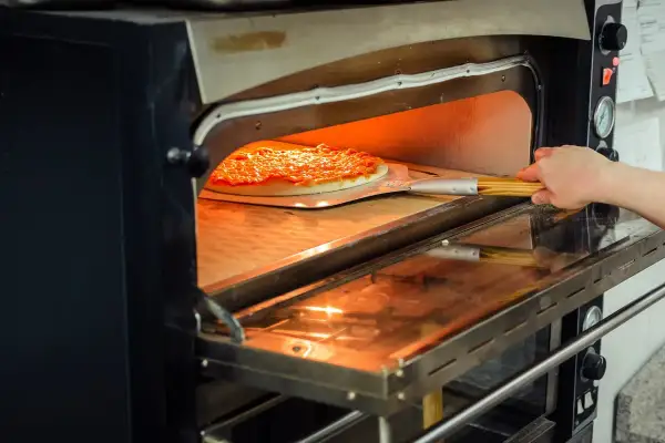 Une main glisse une pizza dans un four à pizza professionnel. La chaleur intense du four est visible, promettant une cuisson parfaite.