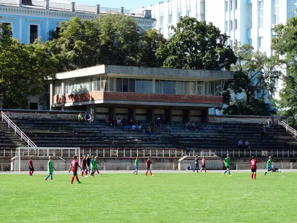 Młodzi piłkarze trenują na boisku. W tle trybuny i budynek, przypominający stadion Dynamo Kijów.