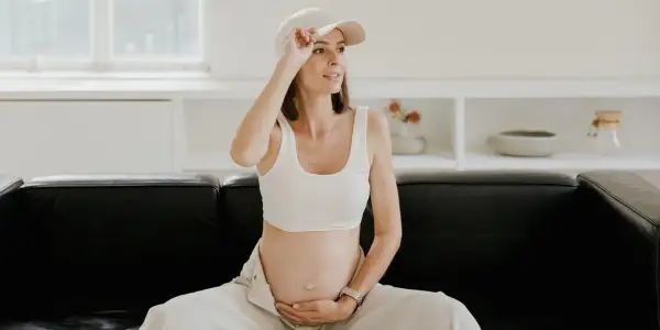 Kobieta w ciąży, w białym topie i czapce, siedzi na czarnej kanapie, czując uderzenia gorąca.