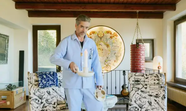 Uomo in pigiama azzurro tiene candela accesa e tazza di caffè, in un ambiente eclettico con opere d'arte e mobili bizzarri. Un'immagine che evoca un momento di riflessione, quasi un rituale mattutino, dove il lavoro incontra il sogno.