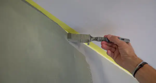 En hand målar en vägg inomhus med en pensel. Gul maskeringstejp skyddar taket.